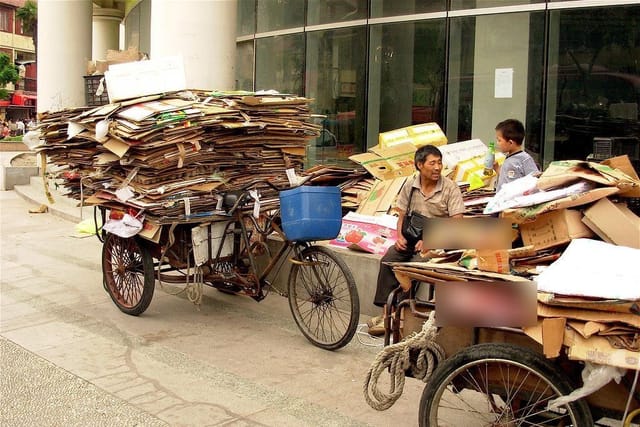 A man rests beside tricycle carts loaded high with flattened cardboard he has collected, with a boy standing nearby.