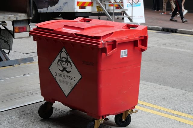 A red wheeled clinical waste bin with a biohazard sign standing beside a collection truck.