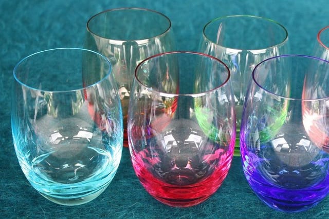 Six stemless drinking glasses tinted blue, pink, green, purple and peach on a teal cloth.