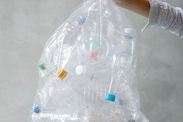 A hand holds up a clear plastic bag stuffed with empty plastic drinks bottles against a grey wall.