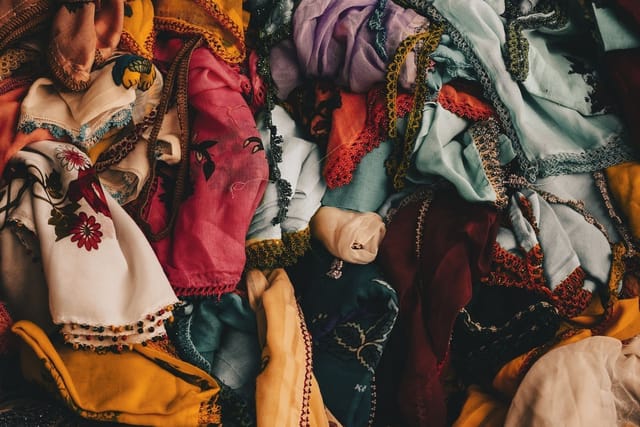 Piles of colourful patterned fabrics and scarves on a market stall, with shoppers behind.