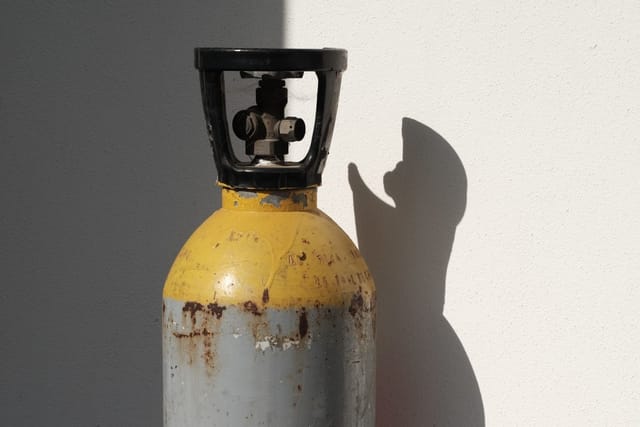 A worn grey and yellow gas cylinder with a black valve guard standing against a white wall.