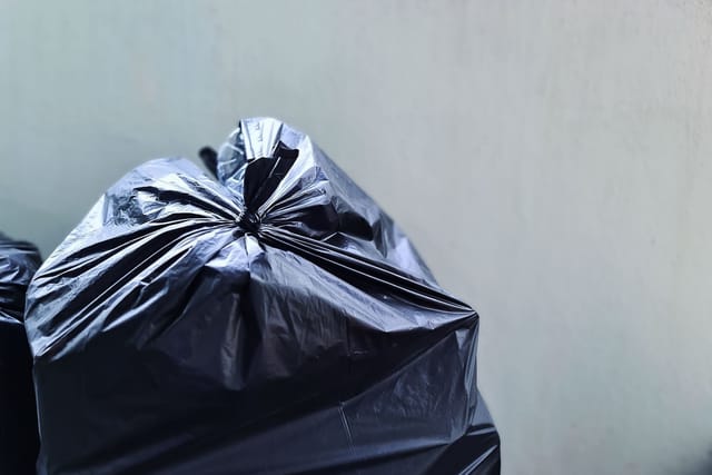 A full black bin bag tied at the top, standing against a grey wall.
