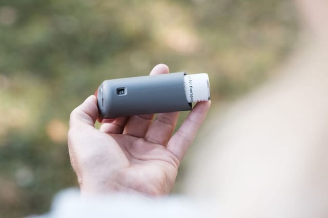 A hand holds a grey asthma inhaler outdoors.