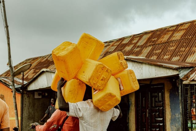 A stack of yellow plastic jerrycans held up in front of buildings with rusty zinc roofs under a grey sky.