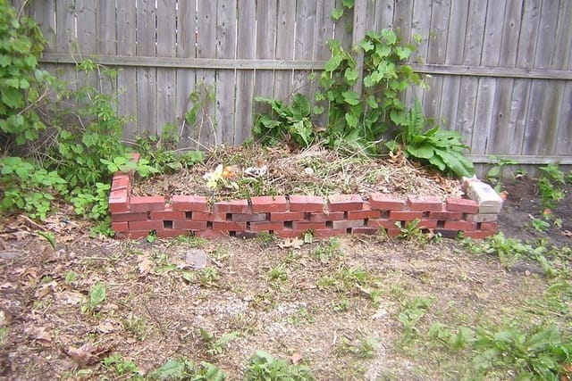 A heap of garden cuttings and leaves inside a low brick edge beside a wooden fence.