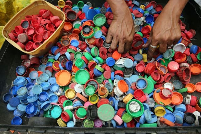 Two hands sort a large tray of loose plastic bottle tops in red, blue, green and orange.