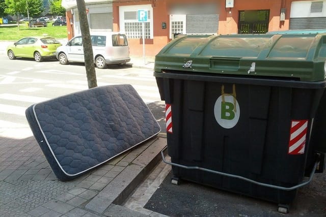 An old mattress left leaning on the pavement beside a large street waste bin.