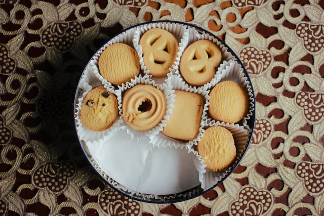An open round metal biscuit tin with butter biscuits in paper cases, on a patterned tablecloth.