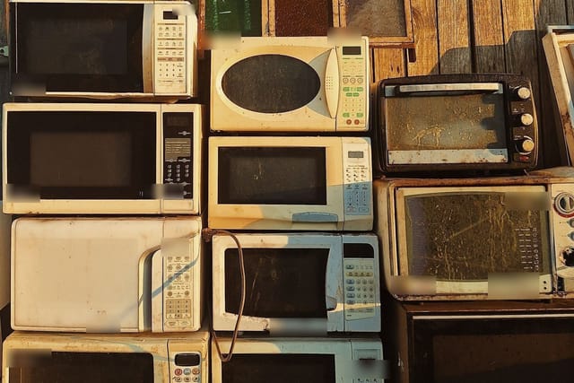 Old microwave ovens and toaster ovens stacked against a wooden wall.