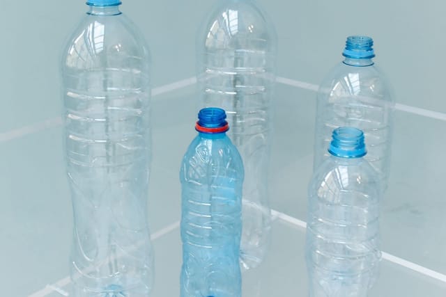 Empty clear plastic bottles with blue caps standing on a pale blue surface, with more crushed bottles on a shelf behind.
