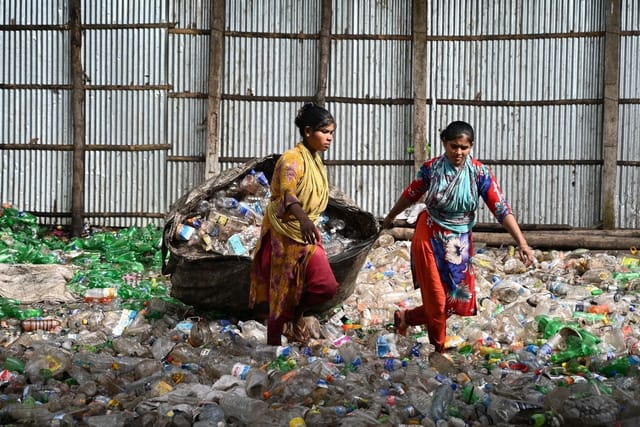 Two women drag a large sack of collected plastic bottles across a yard covered in plastic waste.