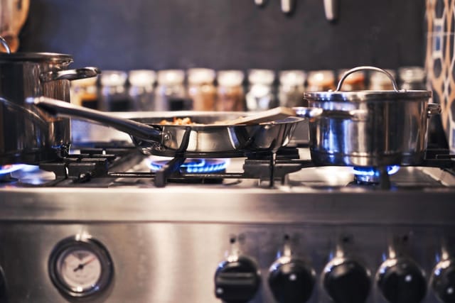 Stainless steel pots and a frying pan on a lit gas cooker in a home kitchen.