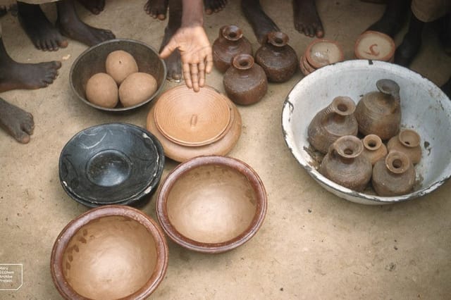 Clay pots, bowls and jars set out on sandy ground, with people standing around them.