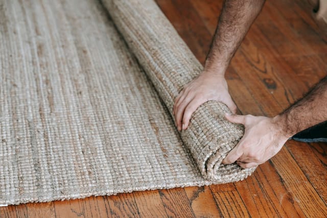 Two hands roll up a woven jute rug on a wooden floor.
