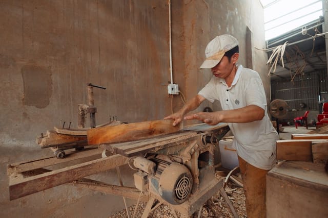 A carpenter in a cap planes a length of timber on a machine in a sawdust-covered workshop.