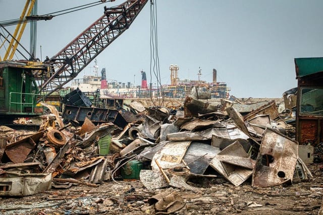 A large heap of rusty scrap metal in a scrapyard with a crane and ships in the background.