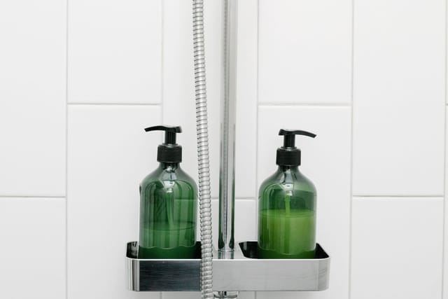 Two green pump bottles of shower gel and shampoo on a metal shower shelf below a hand shower.