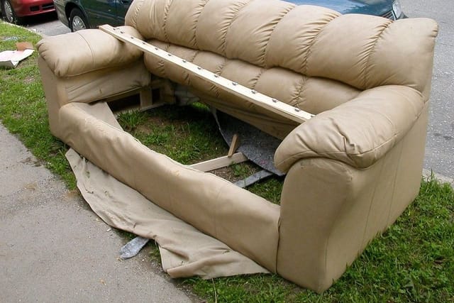 A worn beige sofa left on a grass verge beside a road, with a broken wooden frame on its seat.